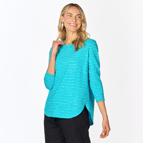 A woman with long blonde hair is wearing the Fresh Produce Clothes Ocean Stripe Catalina Top (Clearance) paired with black pants, smiling and looking to the side against a plain white background.